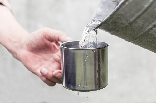 Man’s hand helds a steel mug and a well<br /><br />water is pouring from a bucket